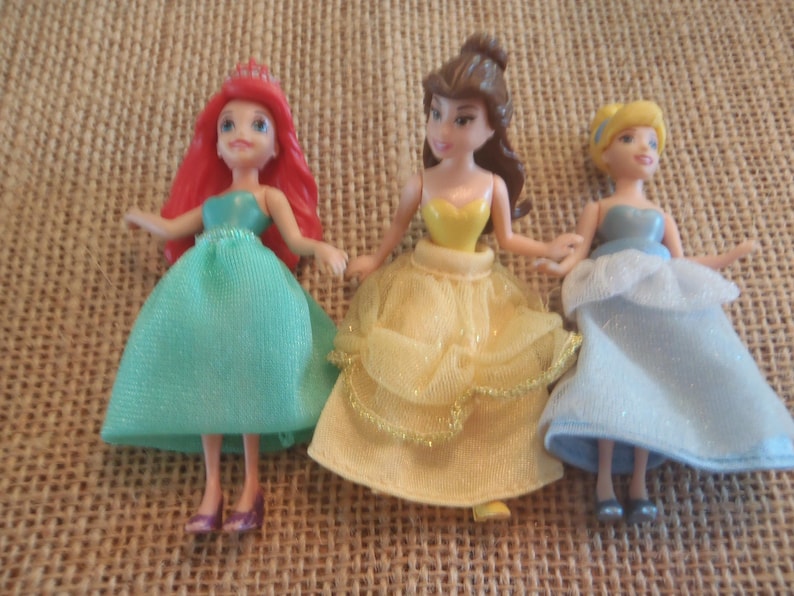 Op de afbeelding: Drie miniatuur prinsessenpoppen. Ariel draagt een zeegroene jurk en heeft rood haar. Belle draagt een gele jurk. Assepoester draagt een lichtblauwe jurk. Alle drie de poppen houden elkaars hand vast en staan samen.
