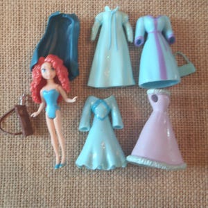 Op de afbeelding: Een pop met rood haar en een blauwe outfit, samen met verschillende miniatuurjurken in blauwe en roze tinten. Een blauwe cape en een bruine koker met een boog zijn ook inbegrepen. De items zijn gerangschikt op een getextureerd, bruin oppervlak.