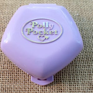 May include: A lavender Polly Pocket compact with the brand name in a silver oval. The compact is closed and has a heart symbol below the brand name. The background is a textured, tan surface.