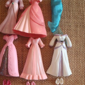 May include: A collection of doll clothes, including several pink and white gowns, a pair of blue pants, and small shoes. The dresses have various designs, some with glitter accents. The items are arranged on a textured, tan surface.