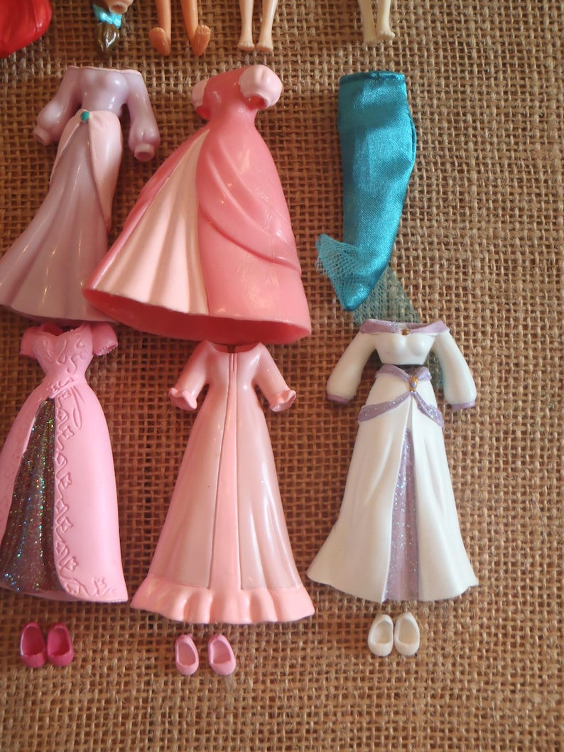 May include: A collection of doll clothes, including several pink and white gowns, a pair of blue pants, and small shoes. The dresses have various designs, some with glitter accents. The items are arranged on a textured, tan surface.