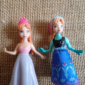 Puede incluir: Dos peque&ntilde;as mu&ntilde;ecas de personajes animados. Una mu&ntilde;eca viste un vestido rosa y blanco con una corona rosa. La otra mu&ntilde;eca viste un vestido azul con un top negro y un tocado azul y rosa. Ambas mu&ntilde;ecas tienen el pelo rojo y ojos azules.