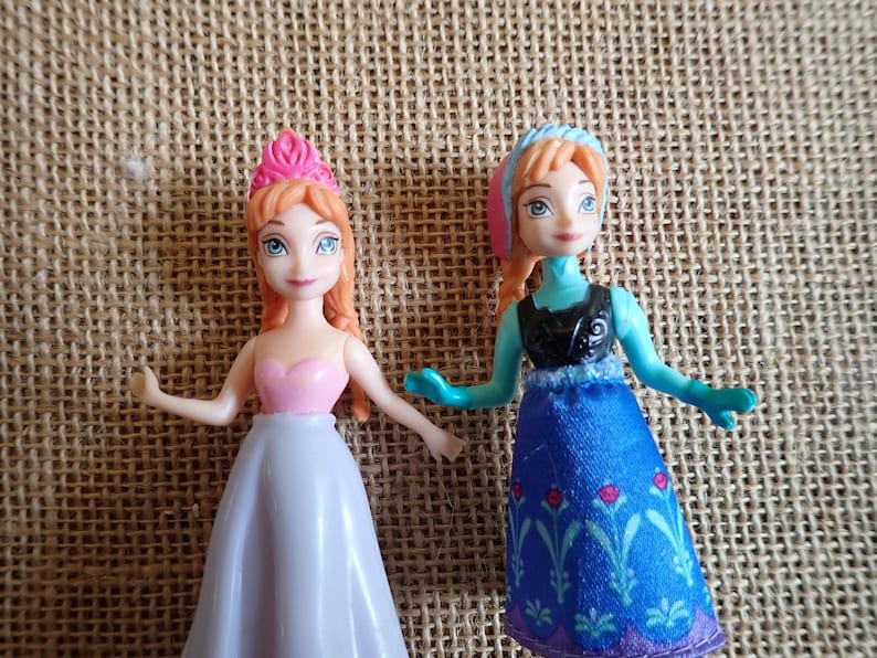 Puede incluir: Dos peque&ntilde;as mu&ntilde;ecas de personajes animados. Una mu&ntilde;eca viste un vestido rosa y blanco con una corona rosa. La otra mu&ntilde;eca viste un vestido azul con un top negro y un tocado azul y rosa. Ambas mu&ntilde;ecas tienen el pelo rojo y ojos azules.