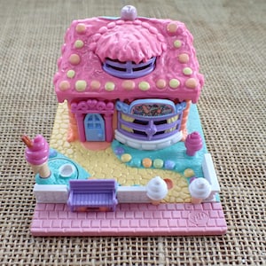 Op de afbeelding: Een miniatuur roze en geel speelgoedhuis met een ijssalon thema. Het huis heeft een roze dak met gele stippen, een blauwe deur en een paarse bank. Decoratieve ijsjes en andere details maken het grillige ontwerp compleet.