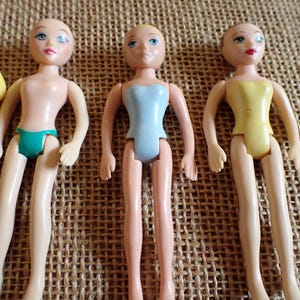 May include: Three small, vintage dolls with painted faces and articulated limbs. One doll wears a teal swimsuit, another a light blue leotard, and the third a yellow leotard. The dolls are on a textured, tan surface.