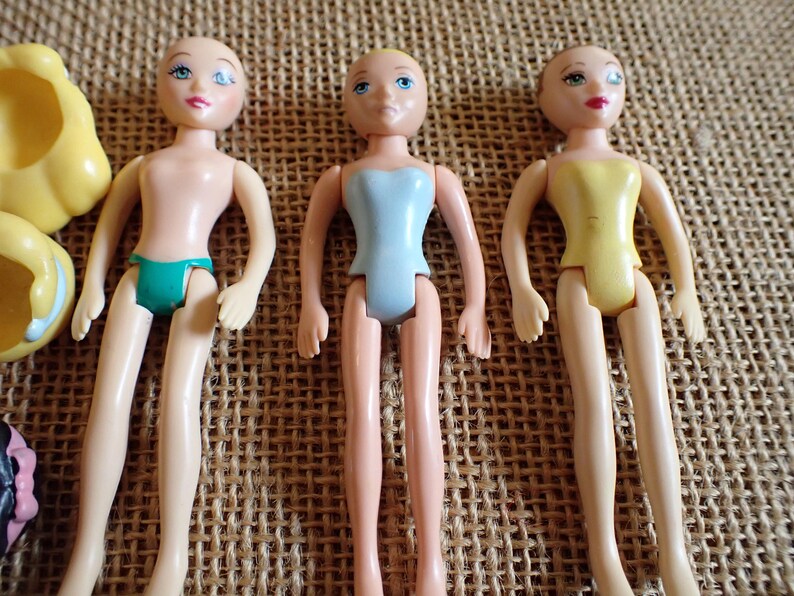 May include: Three small, vintage dolls with painted faces and articulated limbs. One doll wears a teal swimsuit, another a light blue leotard, and the third a yellow leotard. The dolls are on a textured, tan surface.