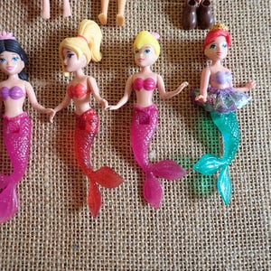 May include: A collection of small mermaid dolls with various hair colors and mermaid tails. The dolls have pink, red, and teal tails, and wear tops in shades of pink and purple. Some dolls have a tutu.