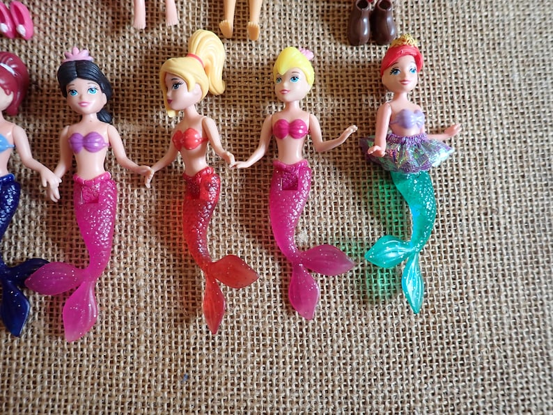 May include: A collection of small mermaid dolls with various hair colors and mermaid tails. The dolls have pink, red, and teal tails, and wear tops in shades of pink and purple. Some dolls have a tutu.