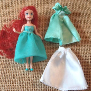 May include: A doll with red hair and a teal dress, along with two additional doll dresses, one teal and one white. The doll is standing on a textured, tan surface. The doll's dress has a sparkly bodice.