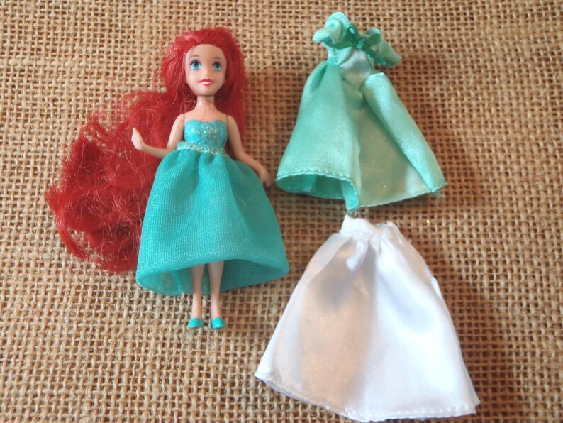 May include: A doll with red hair and a teal dress, along with two additional doll dresses, one teal and one white. The doll is standing on a textured, tan surface. The doll's dress has a sparkly bodice.