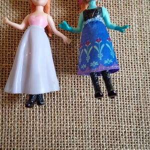 Puede incluir: Dos peque&ntilde;as mu&ntilde;ecas de pl&aacute;stico. Una mu&ntilde;eca lleva un top rosa y una falda blanca. La otra mu&ntilde;eca lleva un vestido azul con estampados florales y botas negras. Las mu&ntilde;ecas est&aacute;n sobre una superficie tejida de color beige.