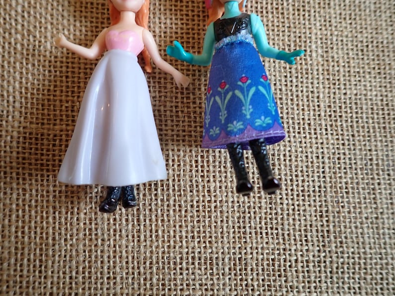 Puede incluir: Dos peque&ntilde;as mu&ntilde;ecas de pl&aacute;stico. Una mu&ntilde;eca lleva un top rosa y una falda blanca. La otra mu&ntilde;eca lleva un vestido azul con estampados florales y botas negras. Las mu&ntilde;ecas est&aacute;n sobre una superficie tejida de color beige.