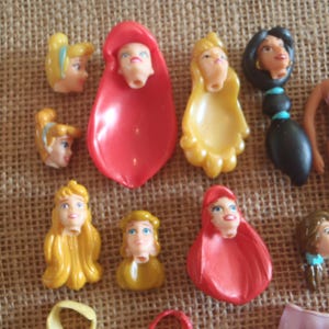 May include: A collection of doll heads and body parts, including several with blonde and brunette hair, and a doll body wearing a pink bikini. The doll heads have painted facial features and are in various colors.