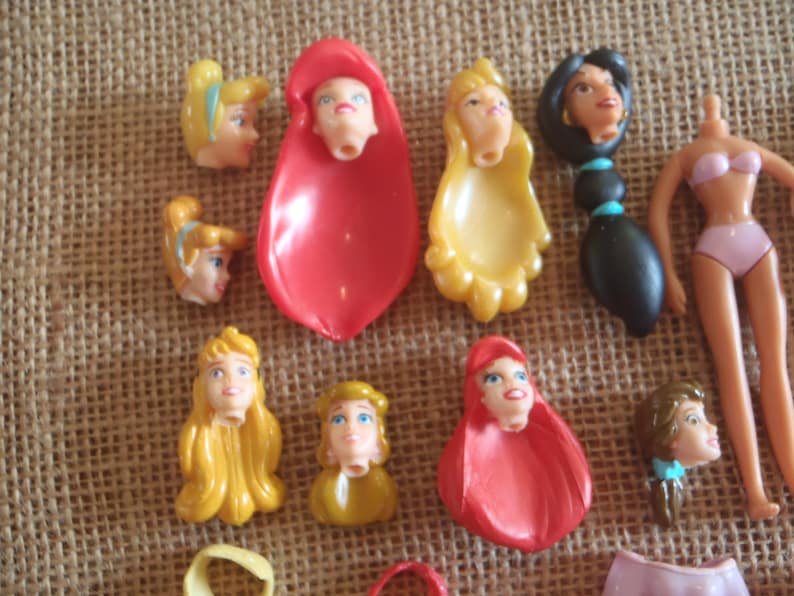 May include: A collection of doll heads and body parts, including several with blonde and brunette hair, and a doll body wearing a pink bikini. The doll heads have painted facial features and are in various colors.