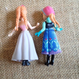 Puede incluir: Dos peque&ntilde;as mu&ntilde;ecas con cabello trenzado naranja. Una mu&ntilde;eca lleva un top rosa y una falda blanca. La otra mu&ntilde;eca lleva un sombrero rosa, guantes azules y un vestido azul con estampado floral. Ambas mu&ntilde;ecas tienen botas negras.