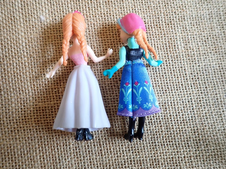 Puede incluir: Dos peque&ntilde;as mu&ntilde;ecas con cabello trenzado naranja. Una mu&ntilde;eca lleva un top rosa y una falda blanca. La otra mu&ntilde;eca lleva un sombrero rosa, guantes azules y un vestido azul con estampado floral. Ambas mu&ntilde;ecas tienen botas negras.