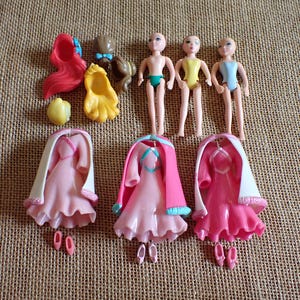 May include: A collection of miniature doll accessories, including three doll figures, dresses, shoes, and hair pieces. The dresses are pink with white capes, and the hair pieces are red, brown, and yellow.