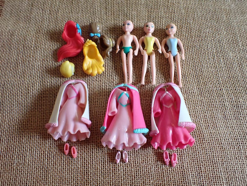 May include: A collection of miniature doll accessories, including three doll figures, dresses, shoes, and hair pieces. The dresses are pink with white capes, and the hair pieces are red, brown, and yellow.