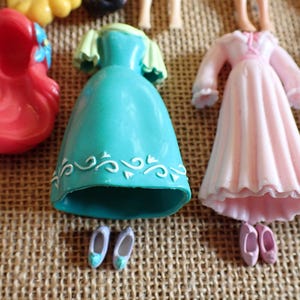 May include: A collection of miniature doll dresses and accessories in various colors. The image includes a red wig, a teal dress with white detailing, a pink dress, and pairs of tiny shoes. The items are arranged on a textured, tan surface.
