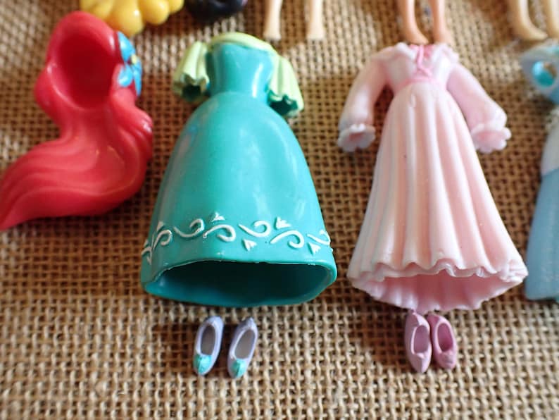 May include: A collection of miniature doll dresses and accessories in various colors. The image includes a red wig, a teal dress with white detailing, a pink dress, and pairs of tiny shoes. The items are arranged on a textured, tan surface.