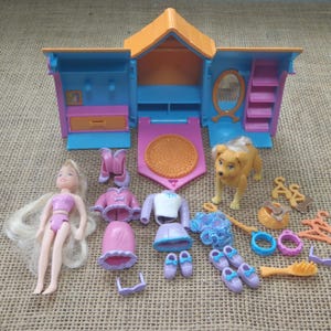 Puede incluir: Un juego de casa de muñecas vintage con una muñeca, una figura de perro y varios accesorios. La casa de muñecas es azul, rosa y naranja. Los accesorios incluyen ropa, zapatos, un peine y perchas. El perro es marrón.