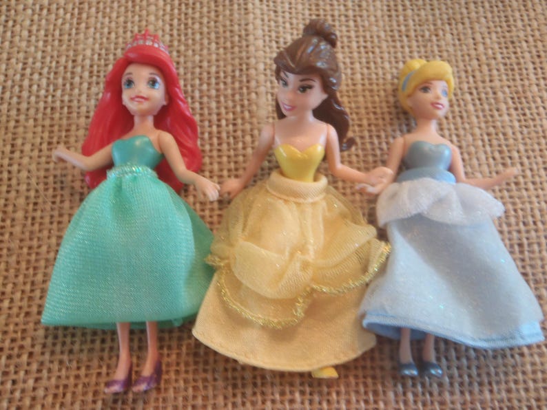 Op de afbeelding: Drie miniatuur prinsessenpoppen. Ariel met rood haar en een groene jurk, Belle in een gele jurk en Assepoester in een blauwe jurk. Elke pop houdt elkaars handen vast.