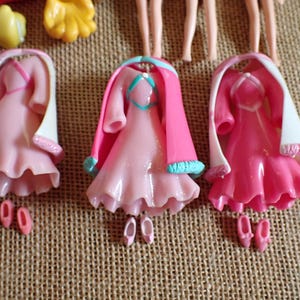 May include: Three pink doll dresses with matching shoes. Each dress has a ruffled skirt, a white shawl, and a pink cape. The dresses are arranged on a textured, tan surface.