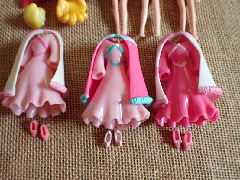 May include: Three pink doll dresses with matching shoes. Each dress has a ruffled skirt, a white shawl, and a pink cape. The dresses are arranged on a textured, tan surface.