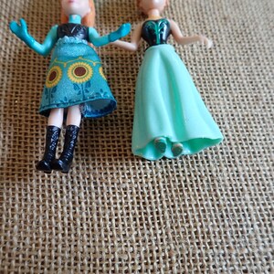 Peut inclure: Deux petites poup&eacute;es en plastique. L'une porte une robe turquoise avec des accents de tournesol jaunes, des bottes noires et des cheveux roux. L'autre porte une robe turquoise et a des cheveux roux. Les poup&eacute;es sont sur une surface textur&eacute;e et beige.