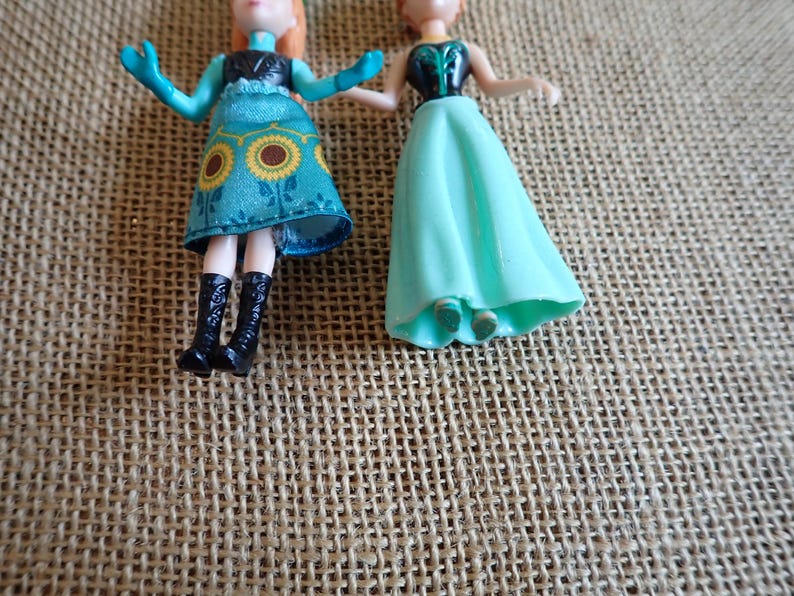 Peut inclure: Deux petites poup&eacute;es en plastique. L'une porte une robe turquoise avec des accents de tournesol jaunes, des bottes noires et des cheveux roux. L'autre porte une robe turquoise et a des cheveux roux. Les poup&eacute;es sont sur une surface textur&eacute;e et beige.