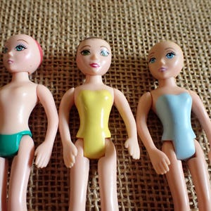 May include: Three small plastic dolls with painted faces and articulated limbs. One doll wears a green swimsuit, another a yellow top, and the third a blue top. The dolls are on a textured, tan surface.