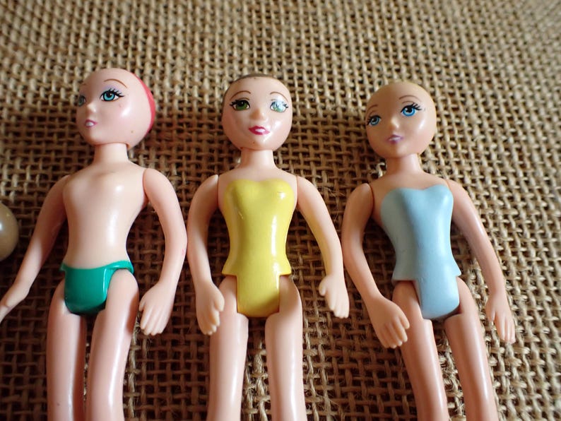 May include: Three small plastic dolls with painted faces and articulated limbs. One doll wears a green swimsuit, another a yellow top, and the third a blue top. The dolls are on a textured, tan surface.