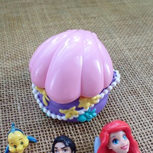 May include: A pink seashell-shaped trinket box with a purple base, decorated with white and yellow details. The box is next to figurines of Ariel, Flounder, and Prince Eric from The Little Mermaid.