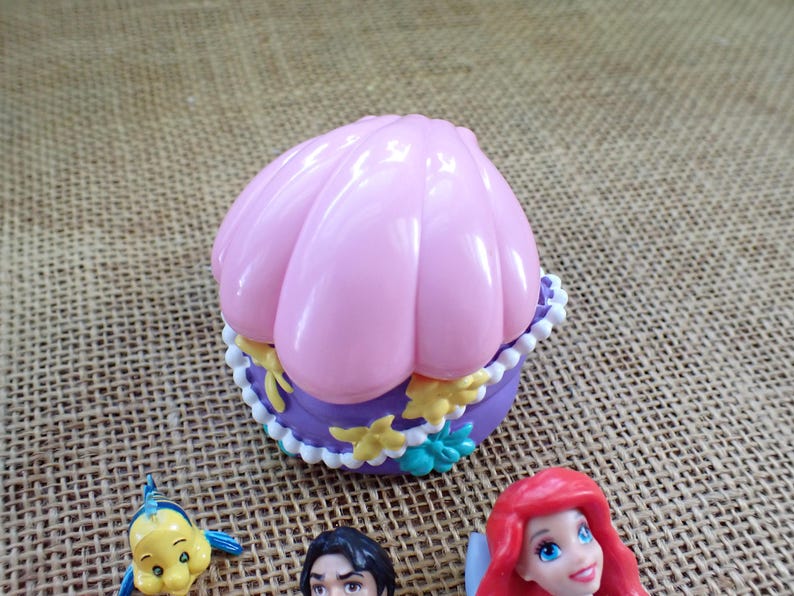 May include: A pink seashell-shaped trinket box with a purple base, decorated with white and yellow details. The box is next to figurines of Ariel, Flounder, and Prince Eric from The Little Mermaid.