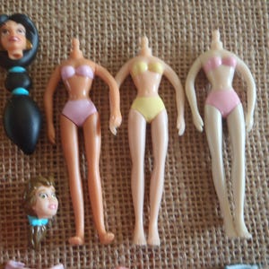 May include: Several doll parts are arranged on a textured surface. The parts include doll torsos in pink and yellow swimwear, and doll heads with long black hair, and blonde hair. The dolls are likely from a children's toy set.