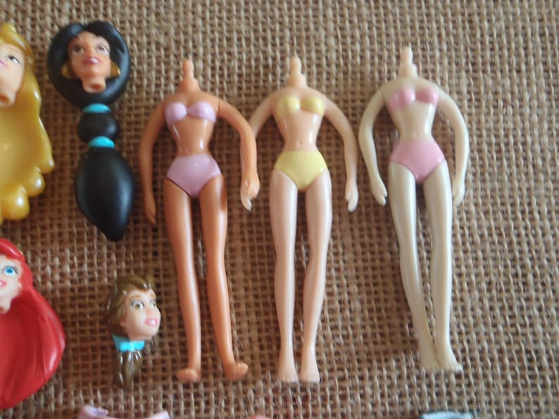 May include: Several doll parts are arranged on a textured surface. The parts include doll torsos in pink and yellow swimwear, and doll heads with long black hair, and blonde hair. The dolls are likely from a children's toy set.