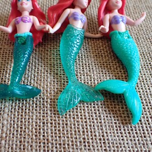 Puede incluir: Tres figuras de sirenas con pelo rojo, tops morados y colas turquesas. Las colas tienen un efecto brillante. Las mu&ntilde;ecas est&aacute;n posadas sobre una superficie texturizada y beige.