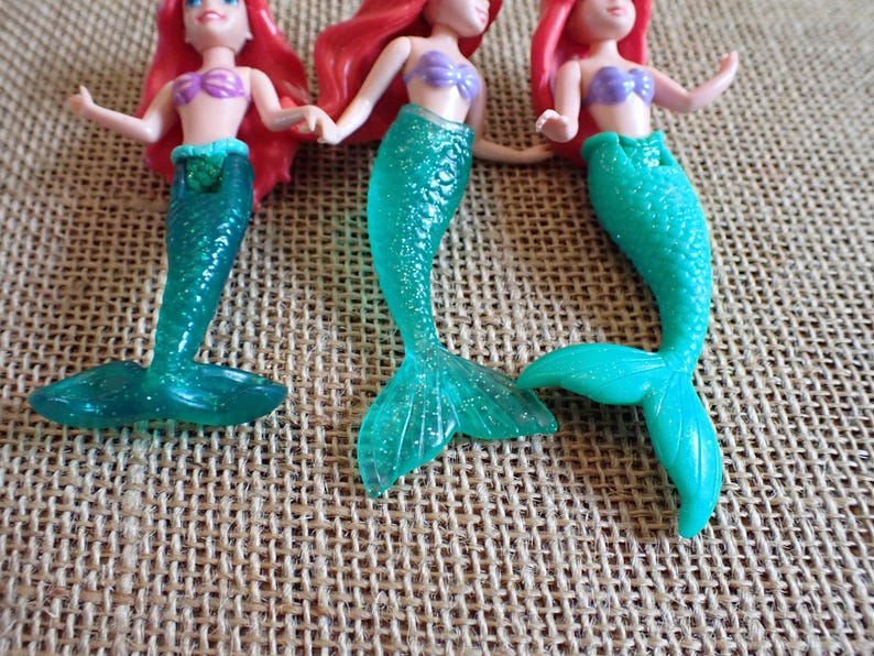 Puede incluir: Tres figuras de sirenas con pelo rojo, tops morados y colas turquesas. Las colas tienen un efecto brillante. Las mu&ntilde;ecas est&aacute;n posadas sobre una superficie texturizada y beige.