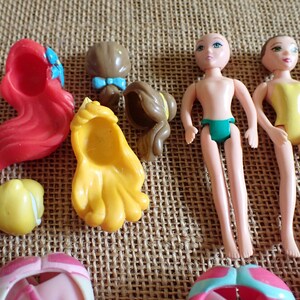 May include: A collection of small plastic doll parts, including doll bodies, hair pieces, and accessories. The hair pieces are in red, yellow, and brown. The doll bodies are nude with green underwear, and one doll is wearing a yellow swimsuit.