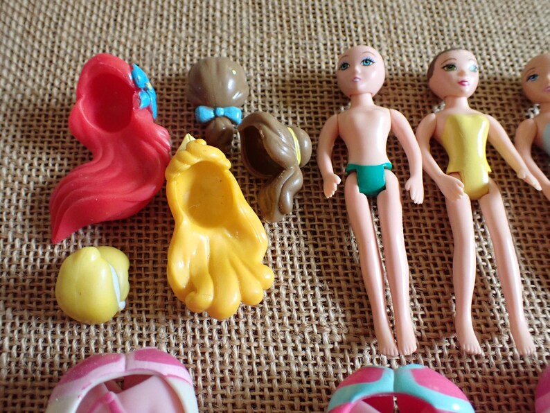 May include: A collection of small plastic doll parts, including doll bodies, hair pieces, and accessories. The hair pieces are in red, yellow, and brown. The doll bodies are nude with green underwear, and one doll is wearing a yellow swimsuit.