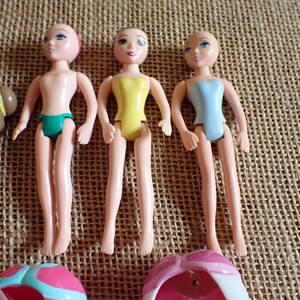 May include: Three small, articulated dolls with painted faces and molded plastic bodies. One doll wears a green swimsuit, another a yellow swimsuit, and the third a light blue swimsuit. The dolls are on a textured, tan surface.
