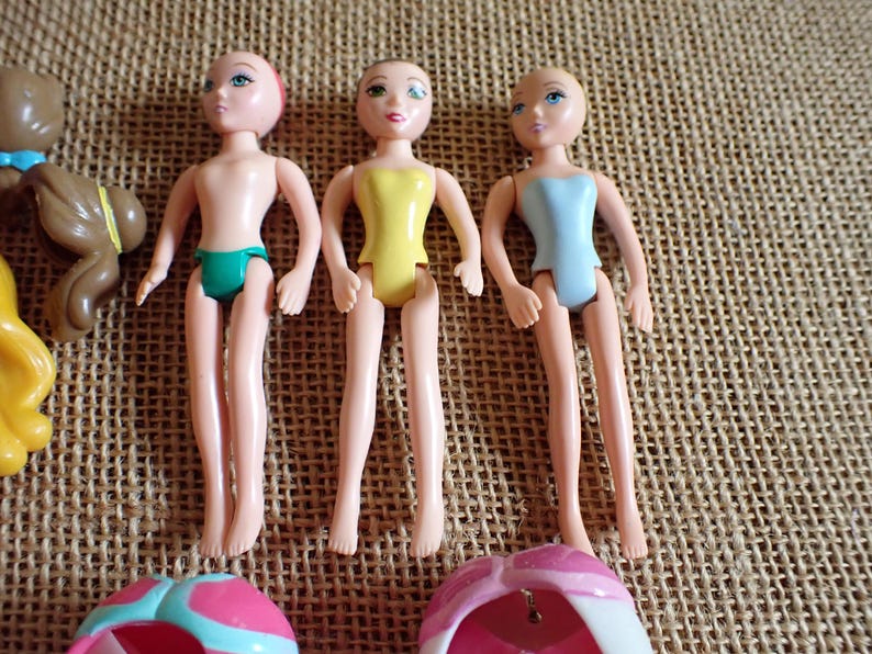 May include: Three small, articulated dolls with painted faces and molded plastic bodies. One doll wears a green swimsuit, another a yellow swimsuit, and the third a light blue swimsuit. The dolls are on a textured, tan surface.