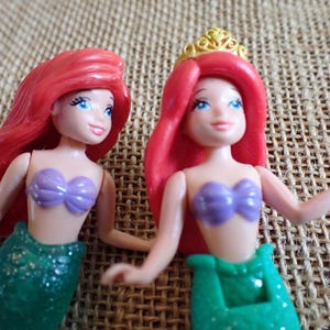 Puede incluir: Dos peque&ntilde;as figuras de sirenas. Cada una tiene cabello rojo, ojos azules y un sujetador de concha p&uacute;rpura. Una lleva una corona dorada. Las colas de sirena son verdes y brillantes. El fondo es una tela texturizada y beige.