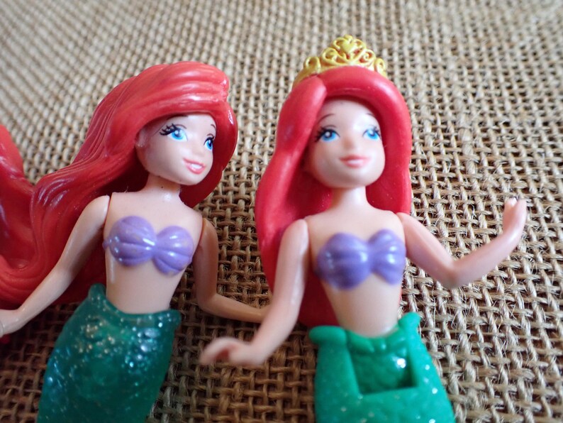 Puede incluir: Dos peque&ntilde;as figuras de sirenas. Cada una tiene cabello rojo, ojos azules y un sujetador de concha p&uacute;rpura. Una lleva una corona dorada. Las colas de sirena son verdes y brillantes. El fondo es una tela texturizada y beige.