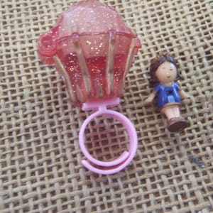 May include: A pink cupcake-shaped ring with gold accents and glitter, next to a small doll wearing a blue dress. The ring is made of plastic and has a round band. The doll has brown hair and brown shoes.