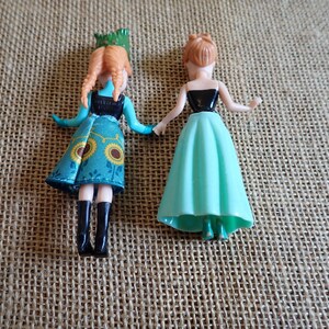 Peut inclure: Deux petites poup&eacute;es sont pr&eacute;sent&eacute;es, chacune avec un design unique. L'une porte une robe turquoise avec des motifs de tournesols et des bottes noires. L'autre poup&eacute;e a un haut noir et une jupe vert clair. Les deux poup&eacute;es ont la peau claire et les cheveux roux.