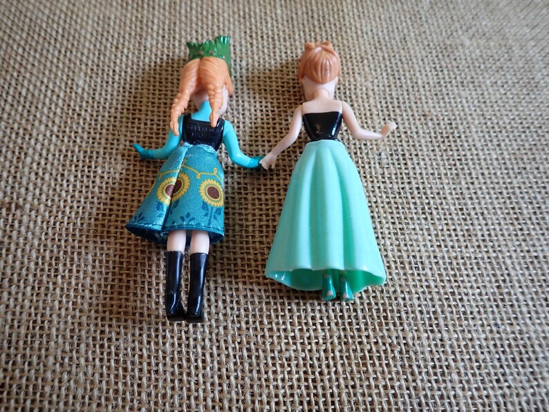 Peut inclure: Deux petites poup&eacute;es sont pr&eacute;sent&eacute;es, chacune avec un design unique. L'une porte une robe turquoise avec des motifs de tournesols et des bottes noires. L'autre poup&eacute;e a un haut noir et une jupe vert clair. Les deux poup&eacute;es ont la peau claire et les cheveux roux.