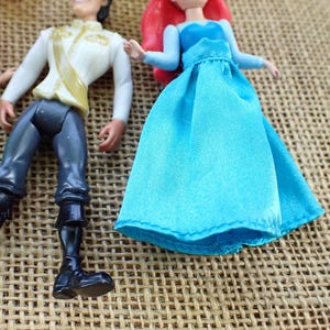 May include: Figurines of Ariel and Prince Eric. Ariel is in a blue dress with red hair. Prince Eric wears a white jacket with gold details and black trousers. The toys are on a textured, tan surface.