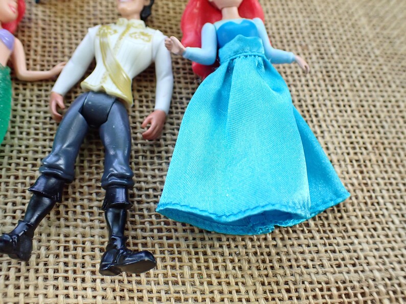 May include: Figurines of Ariel and Prince Eric. Ariel is in a blue dress with red hair. Prince Eric wears a white jacket with gold details and black trousers. The toys are on a textured, tan surface.