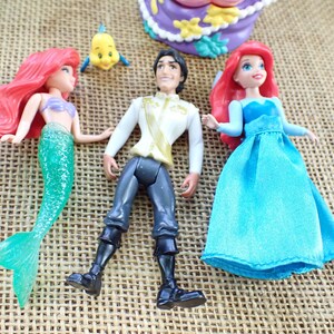 May include: Ariel and Prince Eric dolls from the Disney film, The Little Mermaid. Ariel has a green tail and red hair, and Prince Eric wears a white shirt and black trousers. A small yellow fish is also present.